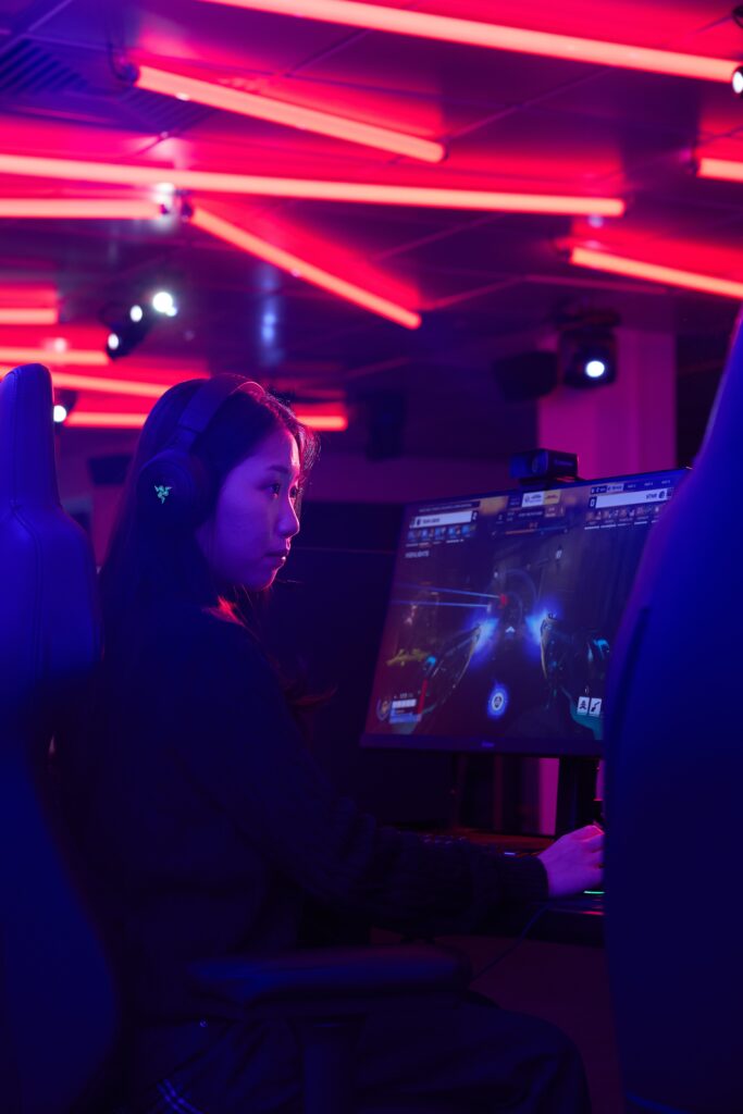 A person wearing headphones plays a video game on a large monitor in a neon-lit gaming room with pink and purple lights.