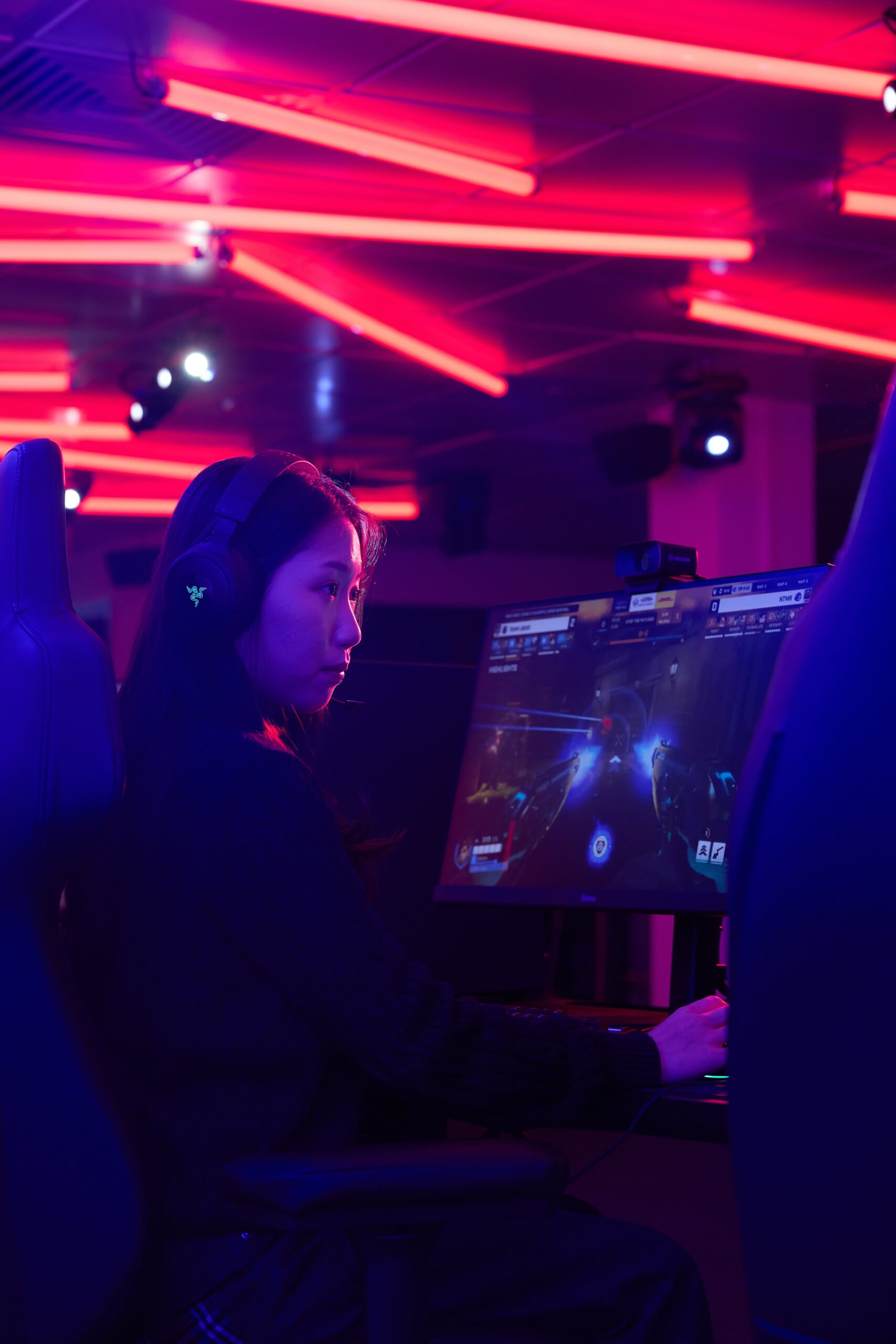 A person wearing headphones plays a video game on a large monitor in a neon-lit gaming room with pink and purple lights.