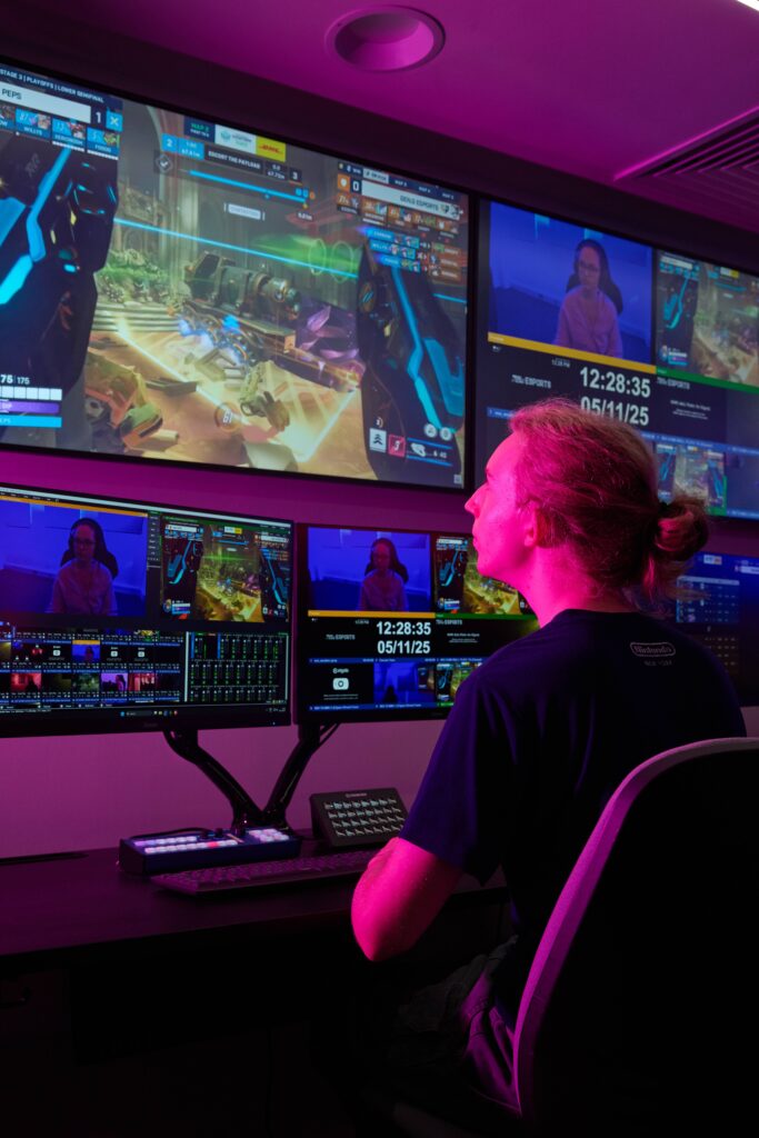 A person sits at a desk with multiple monitors showing a video game and livestream controls, illuminated by purple light in a high-tech gaming studio.