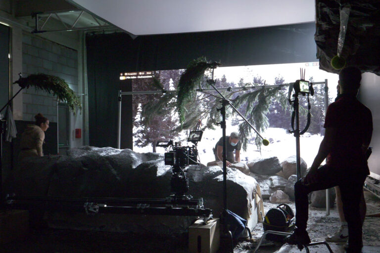 A wide photo of a virtual production film set. Cameras and equipment can be seen in front of a screen displaying a scene with forests and open plains. Several people are busy adjusting the equipment.