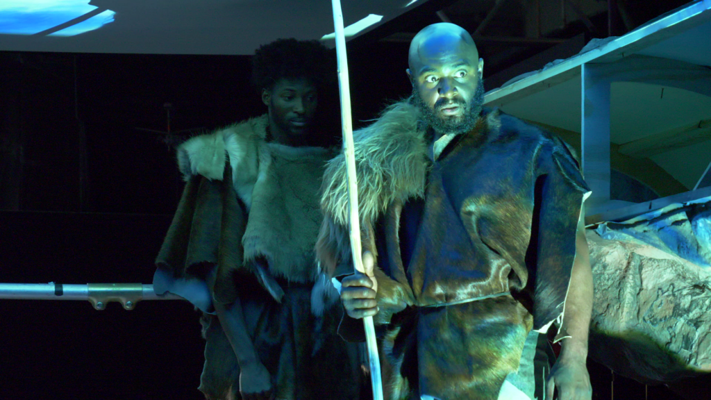 Two people are dressed in fur costumes and holding spears, reminiscent of ancient people from the ice age. A dramatic blue light is shining on them and they have serious expressions.