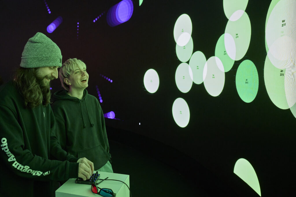 Two people smile as they play with an electronic device that is projecting large circles on a circular screen in front of them