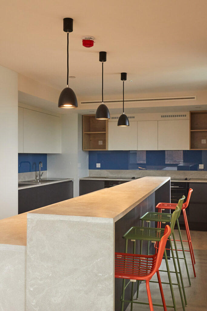 A photograph of the communal kitchen at Duke Street Riverside, Norwich University of the Arts' dedicated student accommodation. In the centre of the image is a long breakfast bar with red and green bar chairs. Around the walls is a modern gloss kitchen with grey and white cabinets