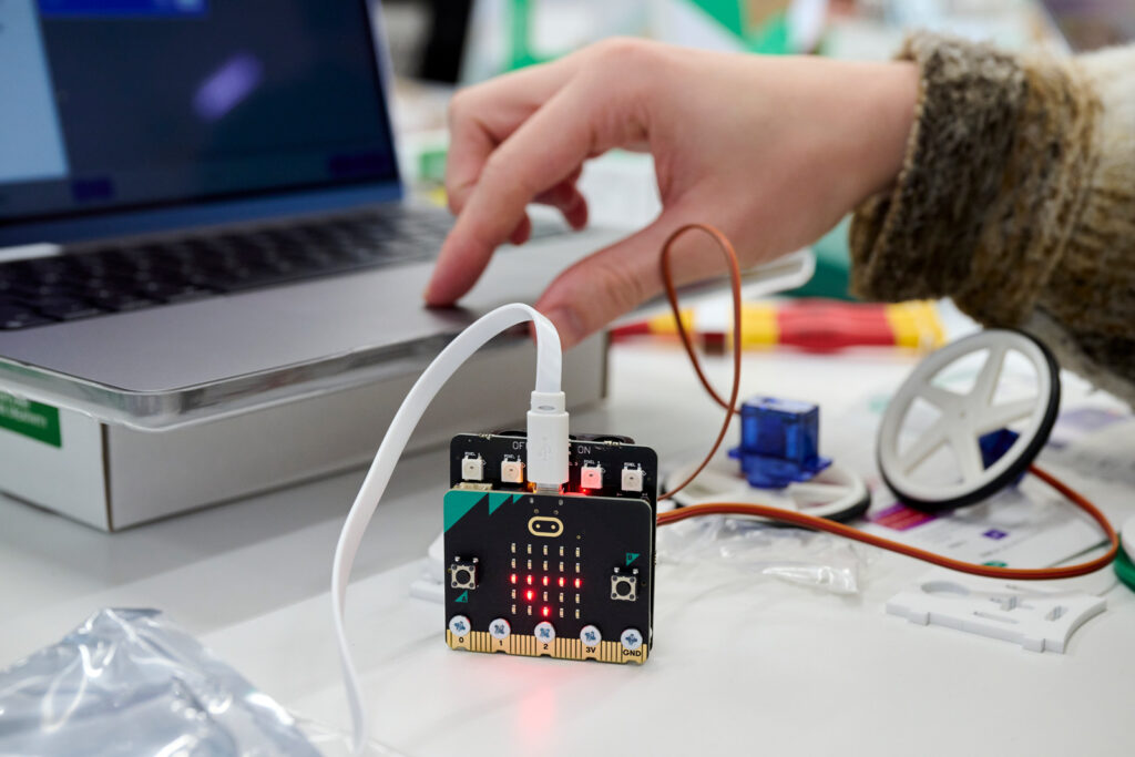 A photo of a motherboard chip standing upright on a table, with some parts lit up. Wires are coming out of the chip. In the background, a person's hand is visible adjusting something on a laptop.