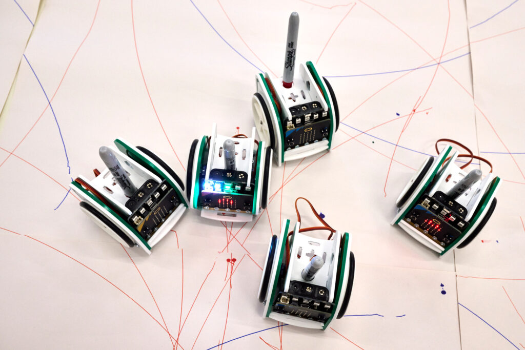 A top-down photo of 5 mini codeable computer robots with sharpie pens mounted inside. On the paper are scribbles where the robot has created marks with the sharpie.