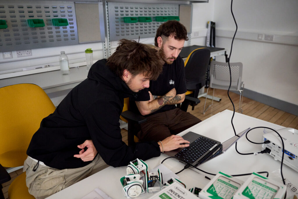 Two students are sat together at a table working on a laptop which is hooked up to a mini codeable computer robot