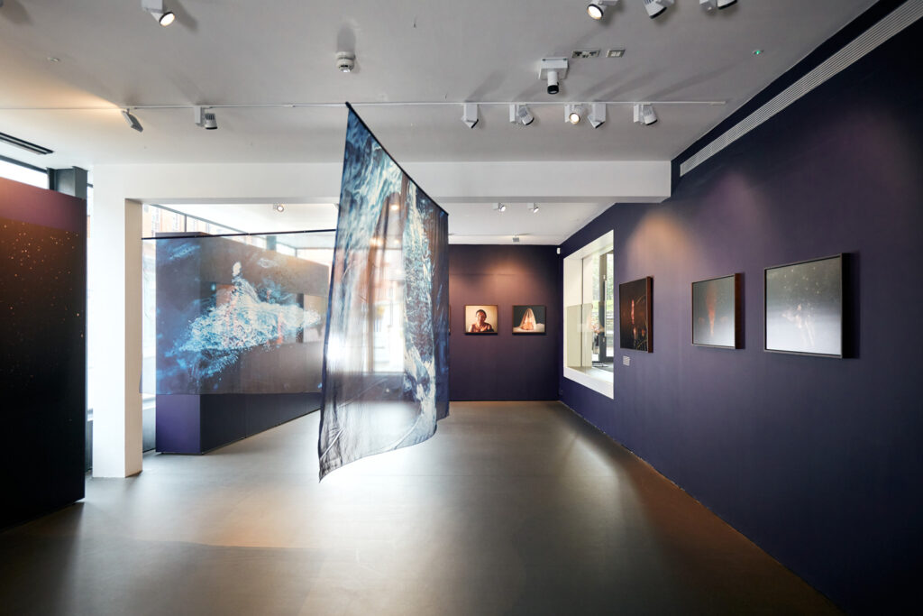 A photograph of the Poulomi Basu exhibition at East Gallery, Norwich. To the right, a wall painted a deep blue has photographs hung upon it. In the middle of the open floor space, a piece of fabric with an image printed on it is suspended from the ceiling