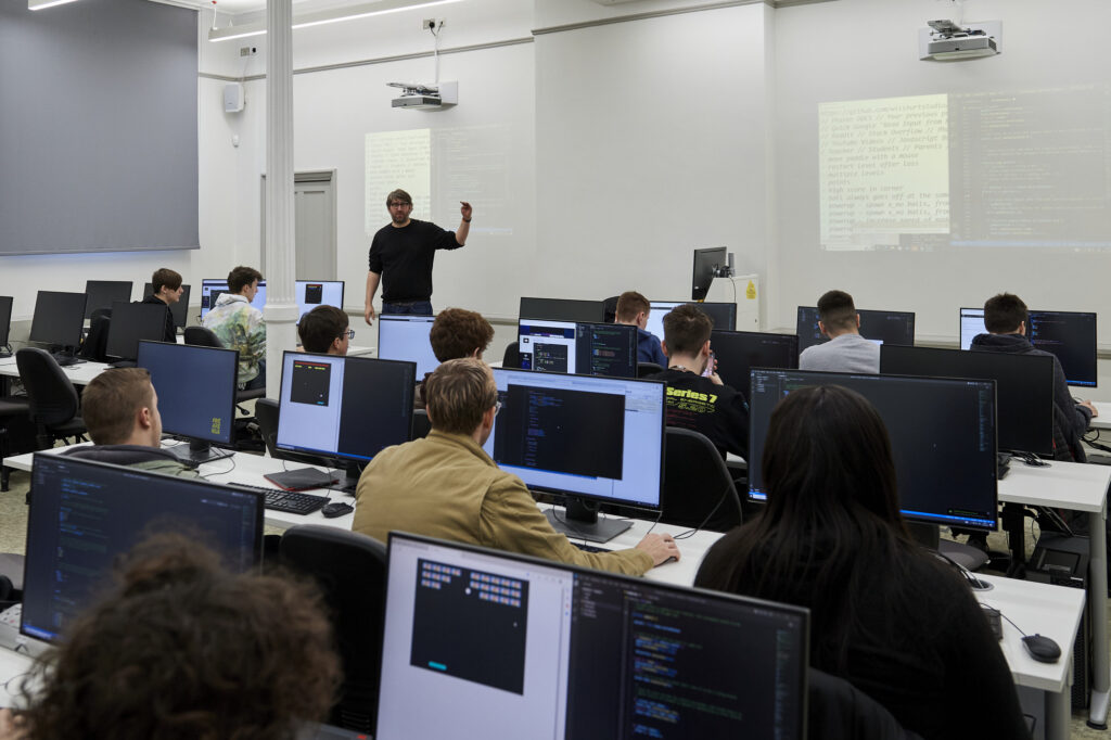 A photo from the back of a computer lab with rows of students at individual workstations. At the front of the room is a lecturer talking to the group, pointing at some projected games coding on the wall