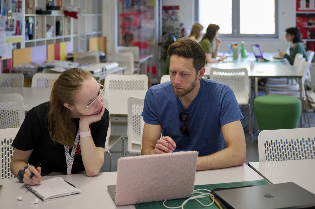 A photograph of a lecturer sat with a student in the graphics studio space, talking over a project on a laptop