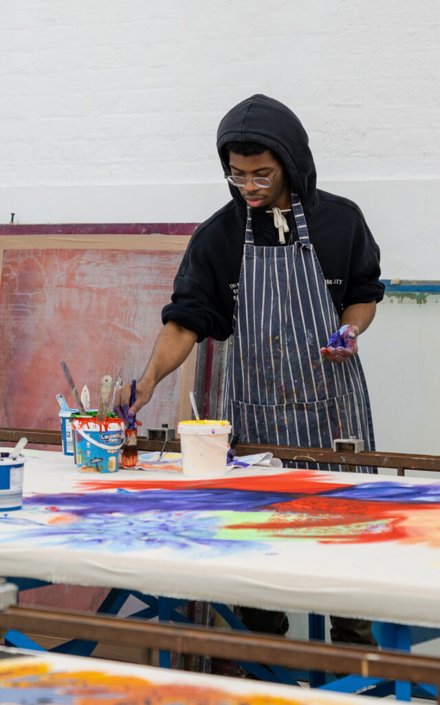 A photo of a student wearing a hoodie and a striped apron, working with coloured dye in the textiles print and dye workshop.