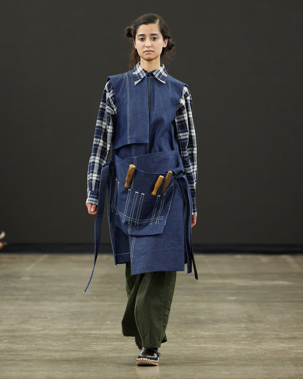 A model walks on a runway at Norwich University of the Arts (NUA), wearing a blue apron-style dress over a plaid shirt, green pants, and black shoes. The apron features attached pockets holding utensils, set against a plain, dark background.