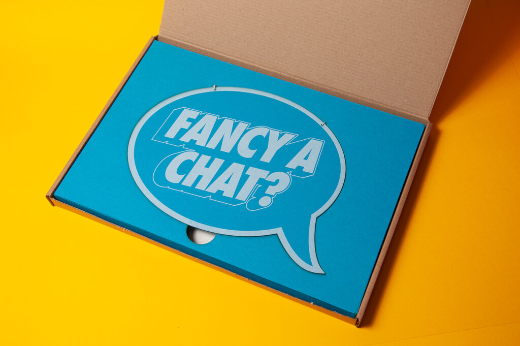 A photograph of a direct mail pack. On a bright yellow background, a cardboard mailer box is open. Inside the box is a bright blue piece of card with the outline of a speech bubble. Inside the speech bubble are the words 'fancy a chat?'.