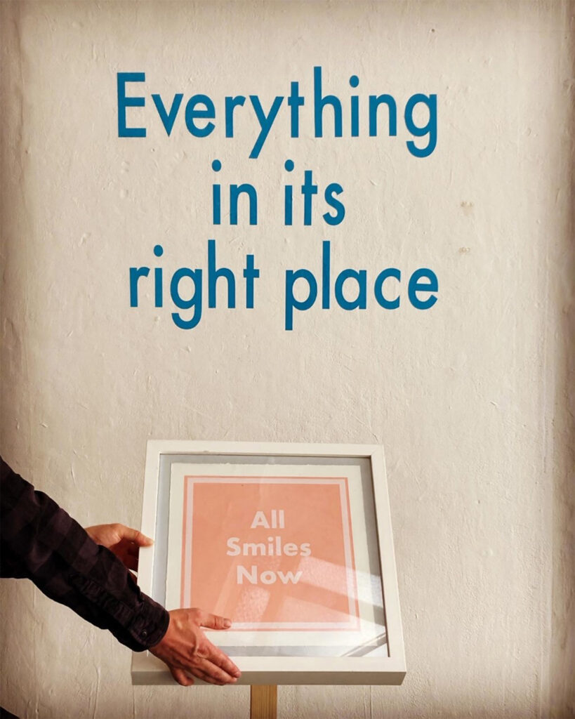 A photograph of a framed piece of typography on a plinth, at an angle facing upwards. The typography reads 'all smiles now'. A pair of hands is reaching to the frame from the left of the image. Above the frame and on the wall are the words 'everything in its right place'