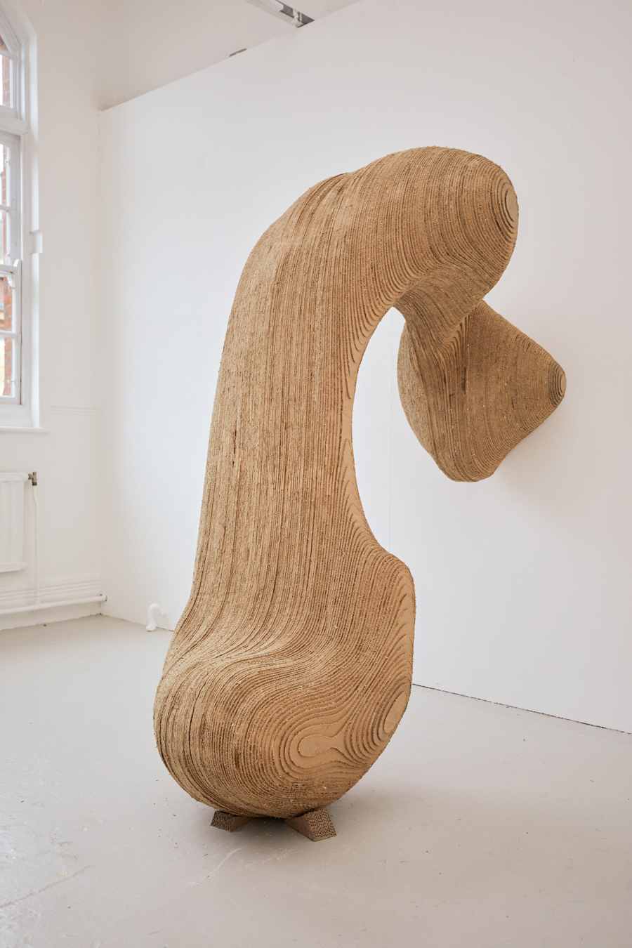 A large, curved sculpture made from cut cardboard. The sculpture resembles a large wooden curved shape, and stands upright against a white wall