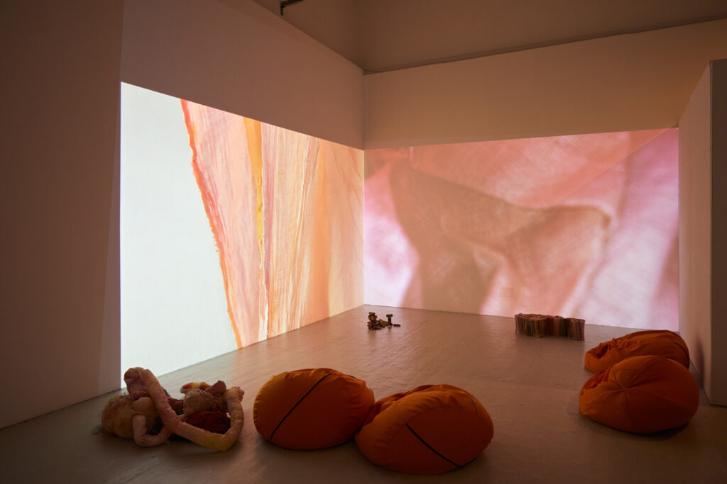 A photograph of an exhibition space with two walls meeting at a corner. Across both walls is a projection of pink, orange and peach coloured fabrics. On the floor in front of the projection are four orange beanbags and sculptured textiles