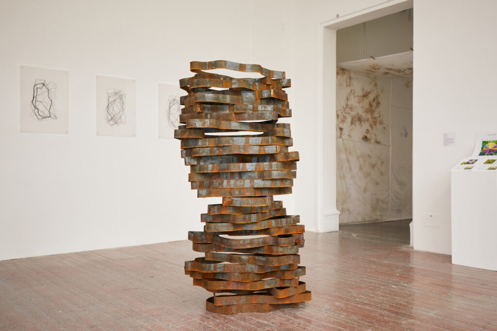 A photograph of a steel stacked sculpture in an exhibition space. Multiple loops of steel, cut into organic wavy patterns resembling thought bubbles, are stacked upon each other.