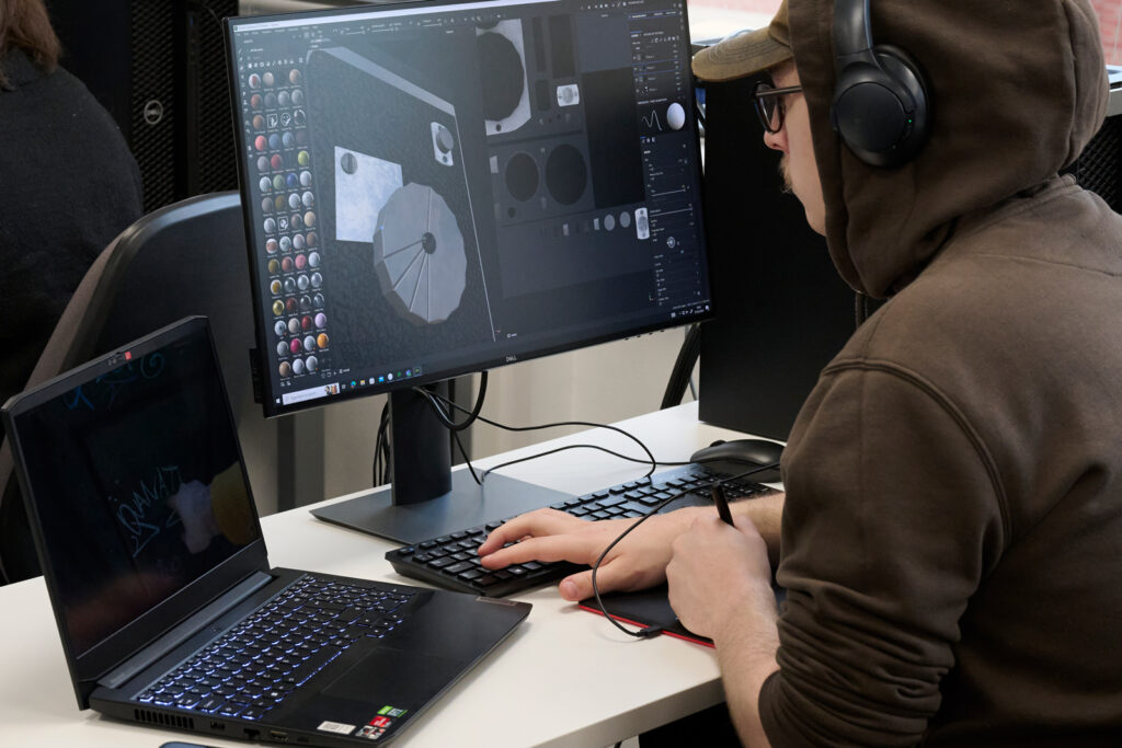 A photograph of a student taken from behind at their side. The student is sat at a desk with a laptop, PC monitor and digital drawing pad.