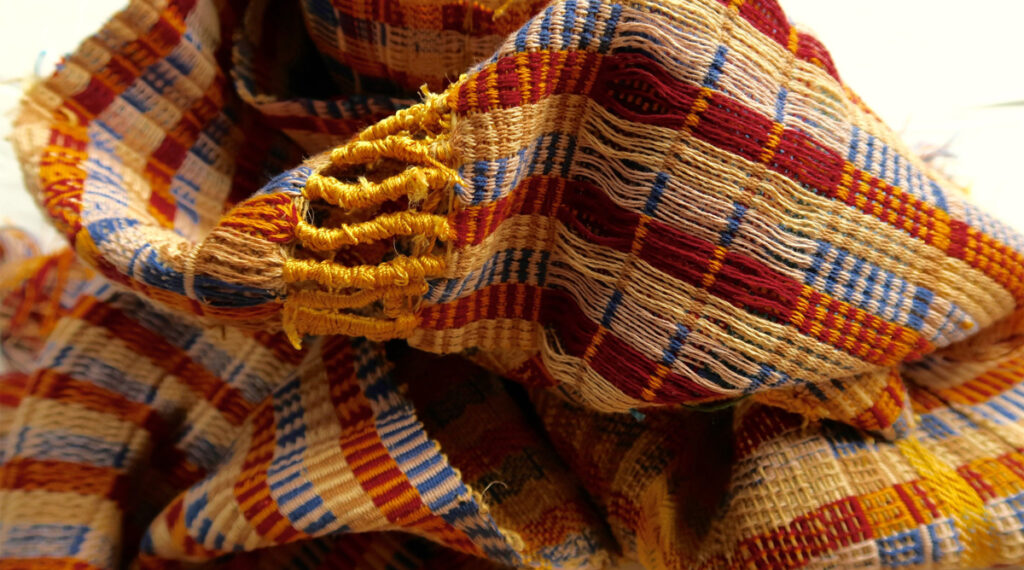 A photograph of woven fabric in a pile. The fabric is made out of areas of tight weaves and loose weaves, i yellow, red, white and blue yarn