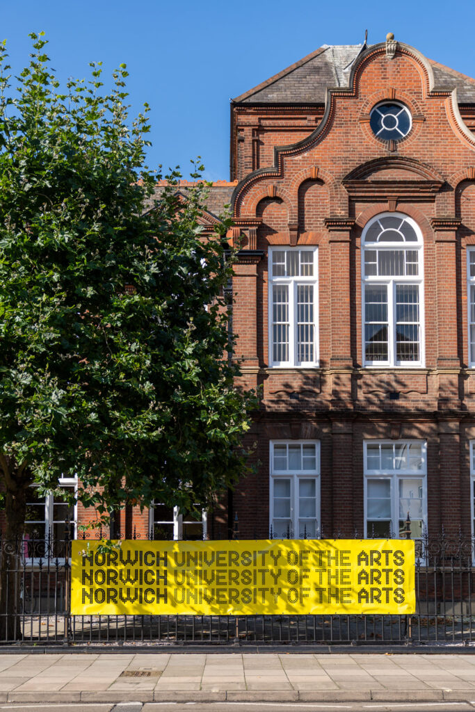 A photo of Norwich University of the Arts' Duke Street building. To the left of the image is a large tree in full bloom. On the iron railings outside the building is a bright yellow banner, with the Norwich University of the Arts logo repeated four times