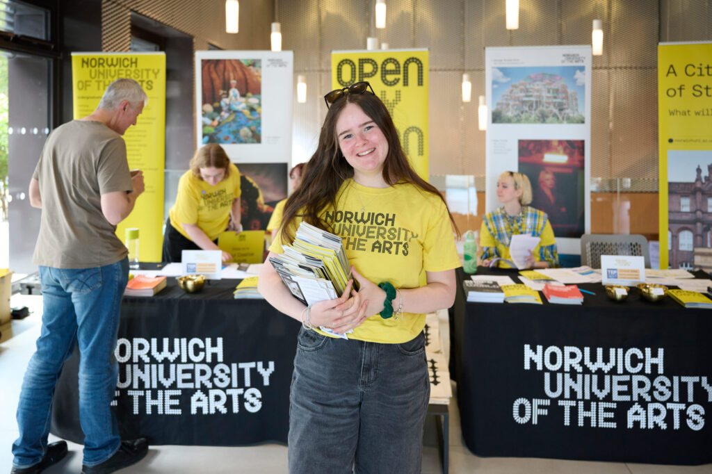 A photo of a female student wearing a bright yellow Norwich University of the Arts t-shirt, holding a stack of Open Day leaflets. Behind the student are two tables with student ambassadors talking to visitors, and some pull up banners of student work and Norwich branding