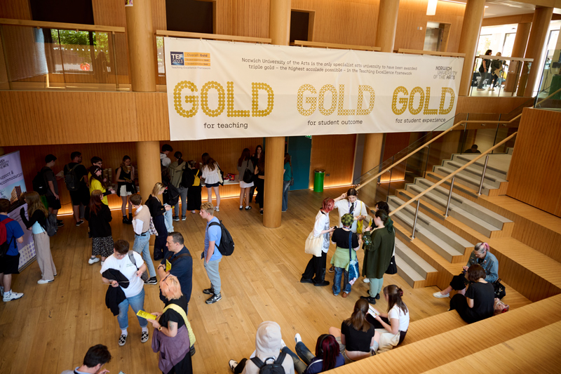 Visitors at an open day at Norwich University of the Arts, photographed in Duke Street Riverside building. Above their heads is a large white banner with 'Gold gold gold' written on it, and information about an award win for the university