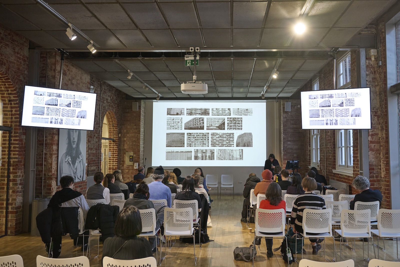People sit facing a presenter in a brick-walled room with two large display screens and a projection of images on the front wall. The audience watches attentively, and the atmosphere is that of a formal lecture or presentation.