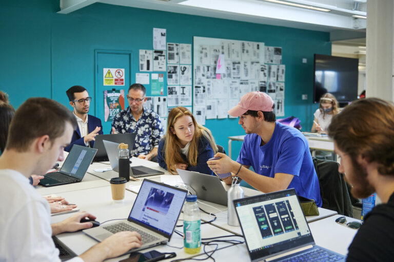 A photograph of students sat around a table as a group, working individually on laptops and discussing projects. The two students in focus are looking at a mobile phone