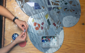 Hands are assembling colorful mosaic tiles on a circular wooden base. Various small pieces of glass are spread around, with a container holding additional tiles. A watch is visible on one wrist. The background is a wooden table.