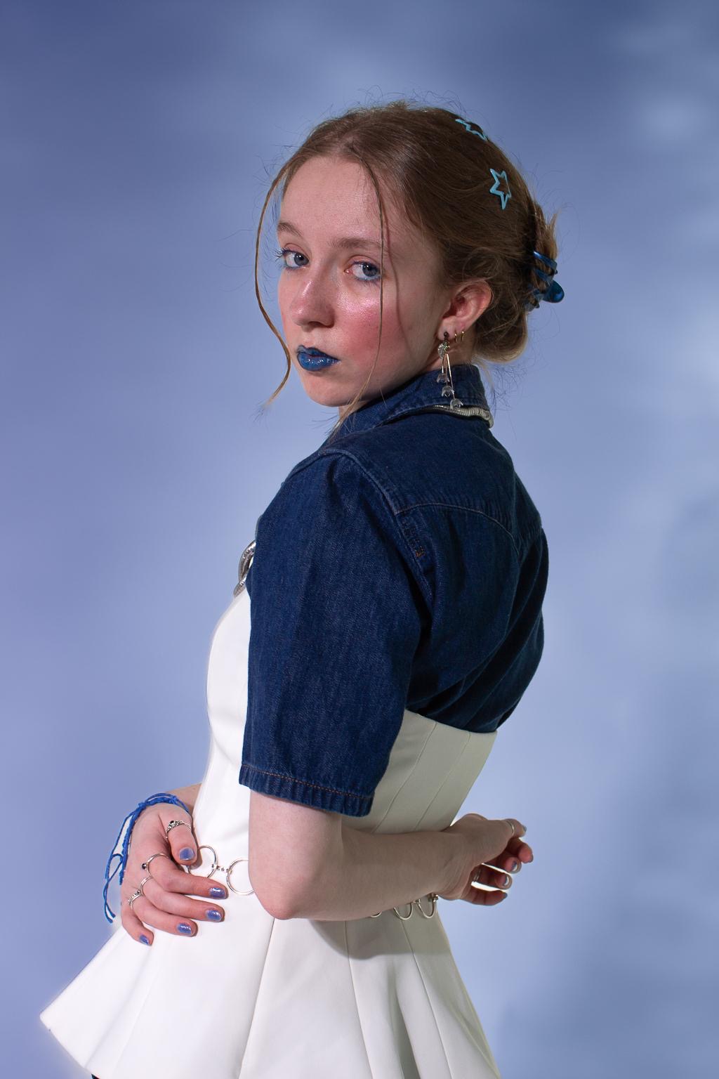 A woman with braided hair and blue accessories stands against a soft blue background. She wears a denim top over a white dress, with matching blue lipstick and nail polish. Her hands are clasped behind her back.