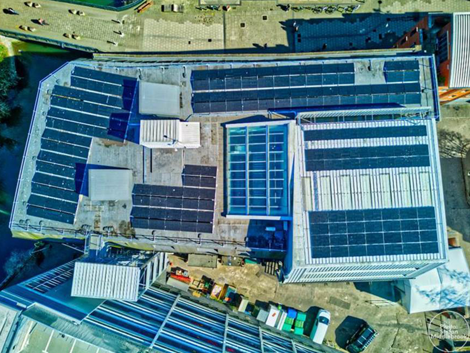 Aerial view of a large industrial building with solar panels covering the rooftop. Surrounding the building are parked cars and other structures. The photo is taken in daylight, highlighting the pattern and layout of the panels.