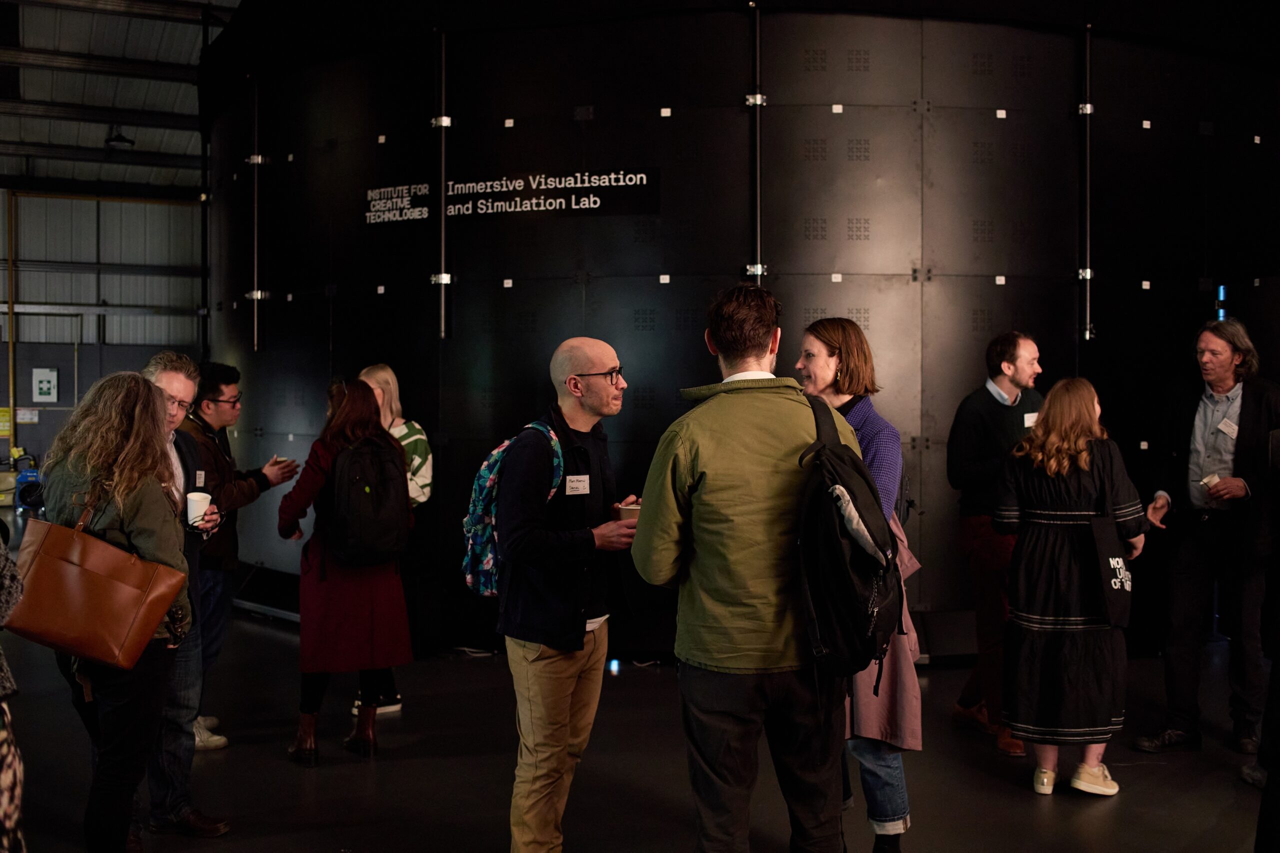 A group of people engaged in conversation inside a dimly lit room with a large black immersive visualization and simulation lab structure in the background. Some are holding drinks, and they appear to be at an informal event or gathering.