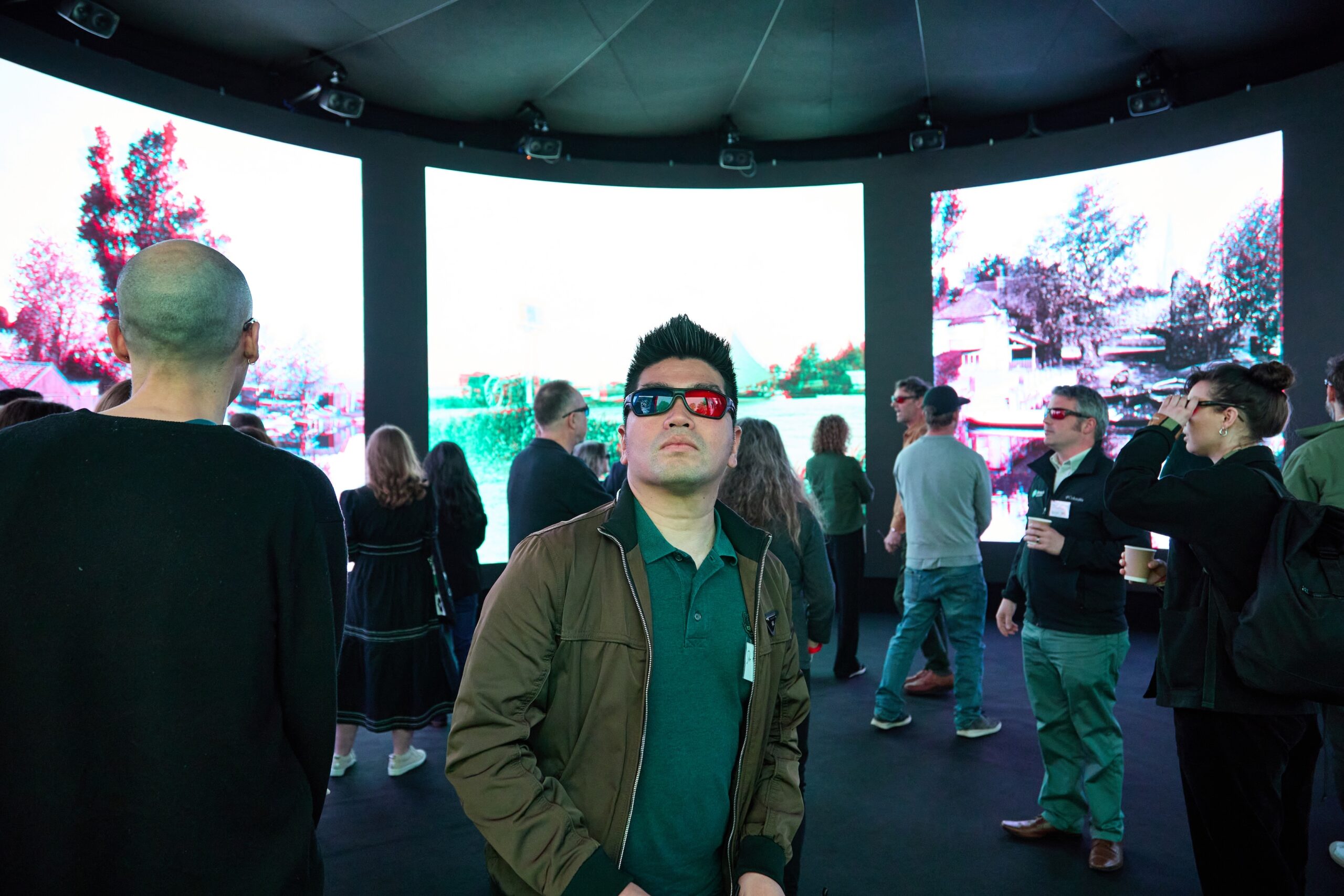 A group of people wearing 3D glasses are inside a dark room with large, bright screens displaying scenes. A person in the foreground wears a green jacket and looks up at the screens.