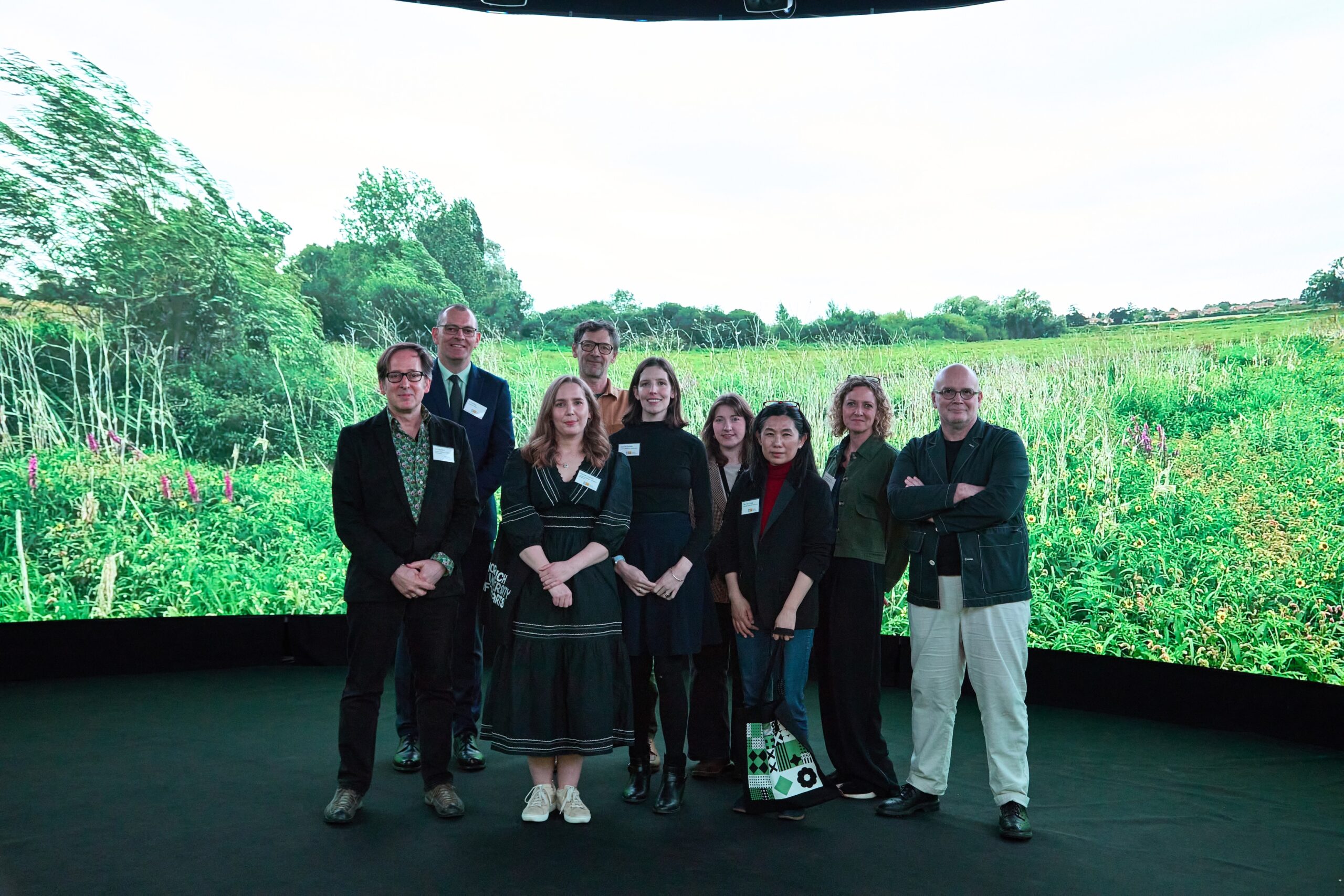 A group of nine people standing together indoors in front of a large, curved screen displaying a lush green meadow with trees. The individuals are in casual to semi-formal attire and are posing for a group photo.