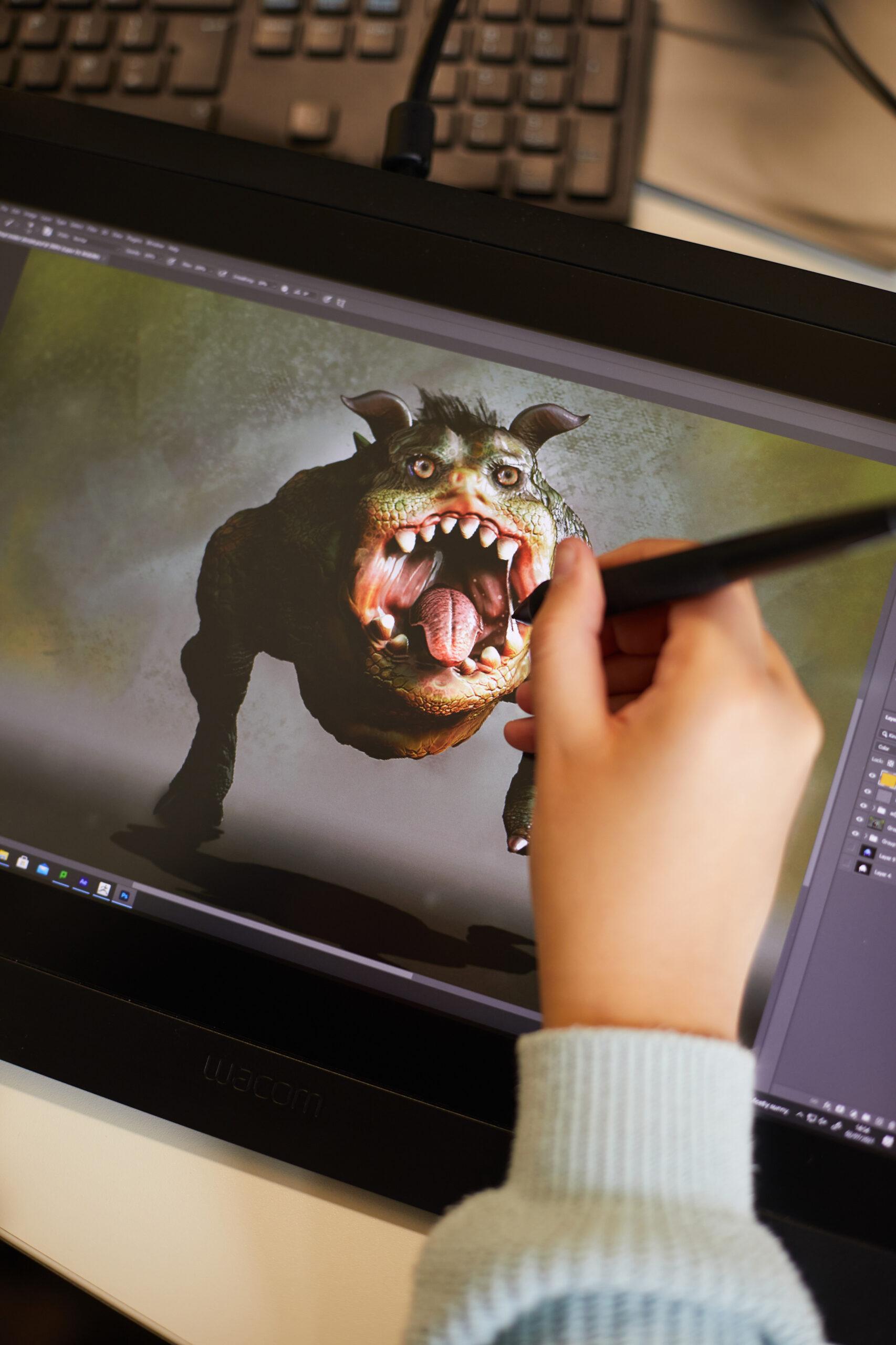 A person uses a stylus on a graphics tablet to create a digital image of a green, monstrous creature with large teeth and horns. The tablet displays detailed artwork, and a computer keyboard is visible in the background.