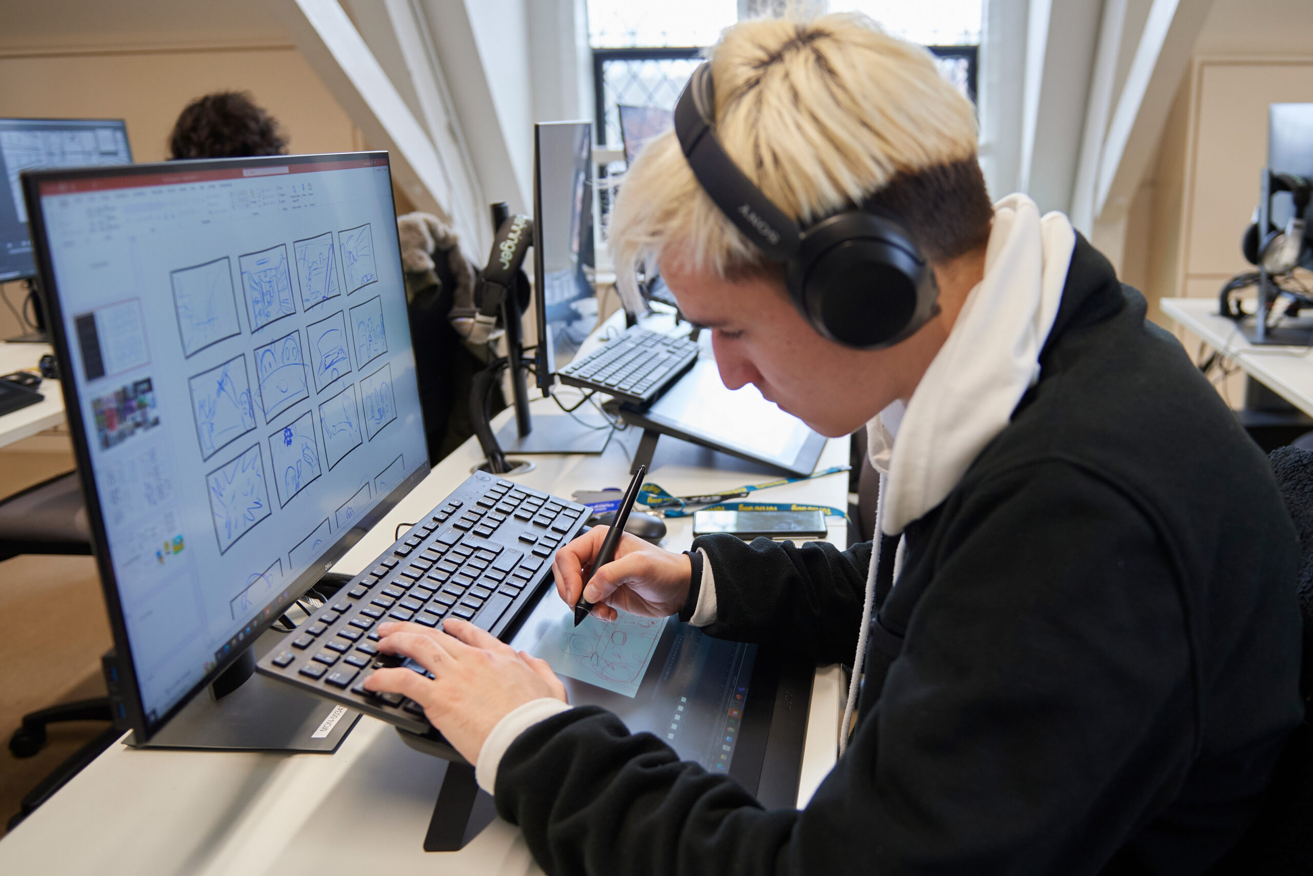 A person with headphones is working at a computer with a dual-screen setup. They are drawing on a tablet display, focusing on creating illustrations. Various sketches are visible on the screen. Office setting with other computers in the background.