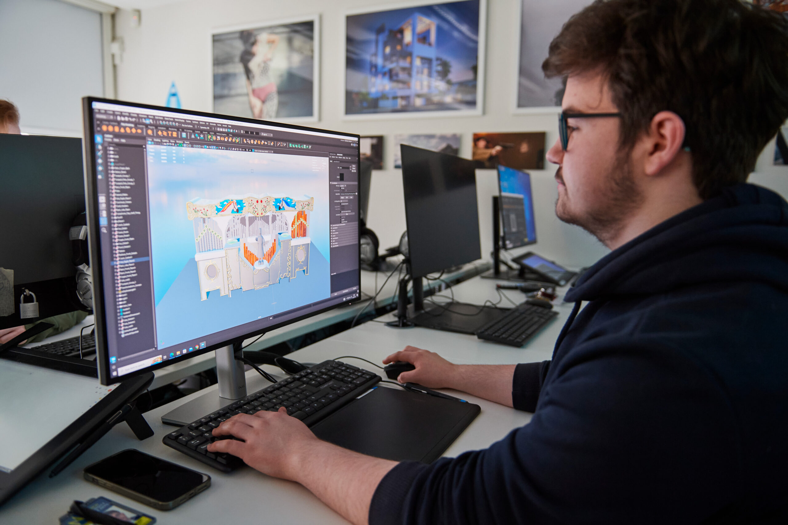 A person using 3D modeling software on a computer, focusing on a detailed design of an architectural structure. The workspace includes multiple monitors and framed architectural images on the wall.