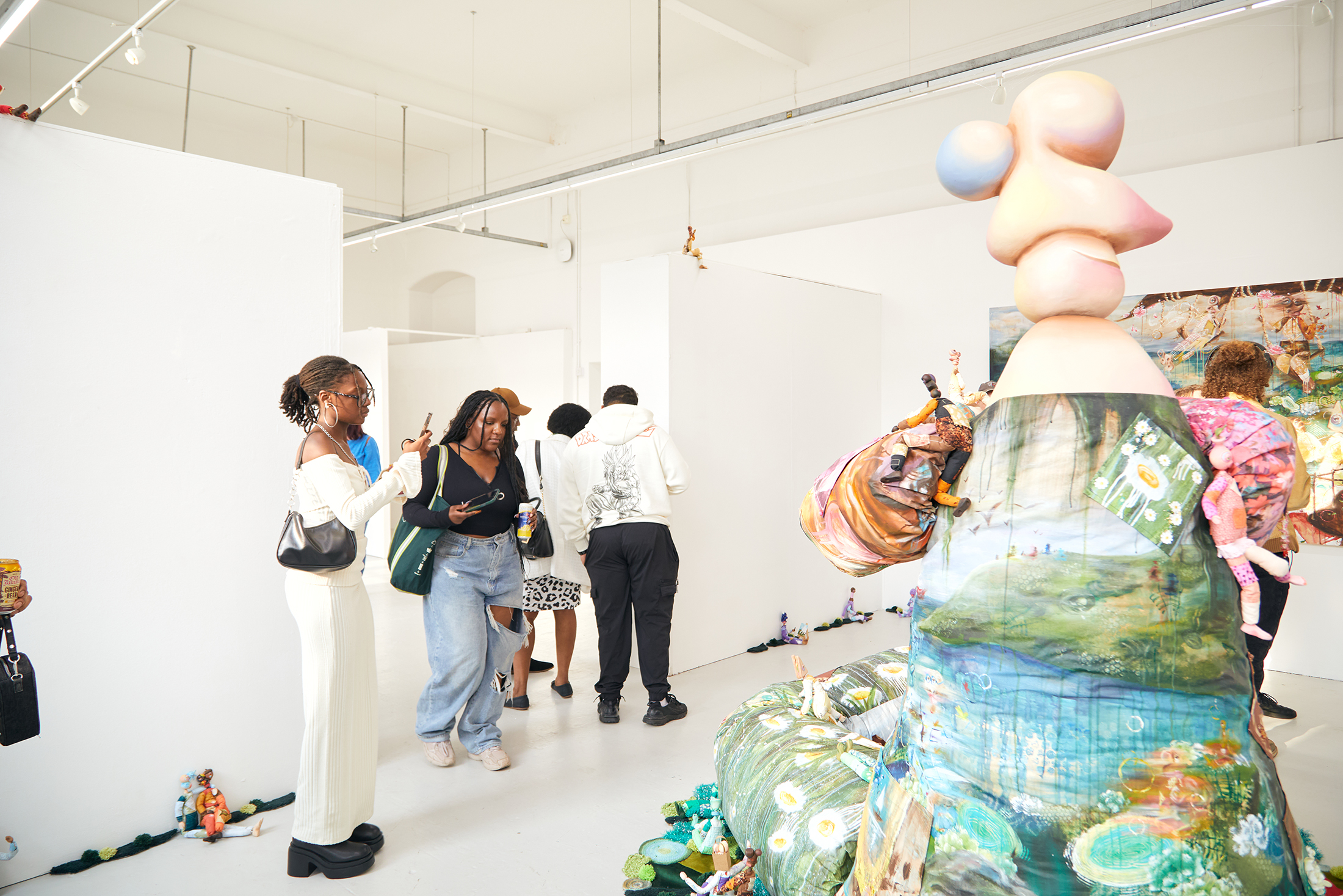 People view colorful contemporary art sculptures and paintings in a bright, modern gallery space. One large abstract sculpture is in the foreground, while several visitors observe the artworks and interact with each other.
