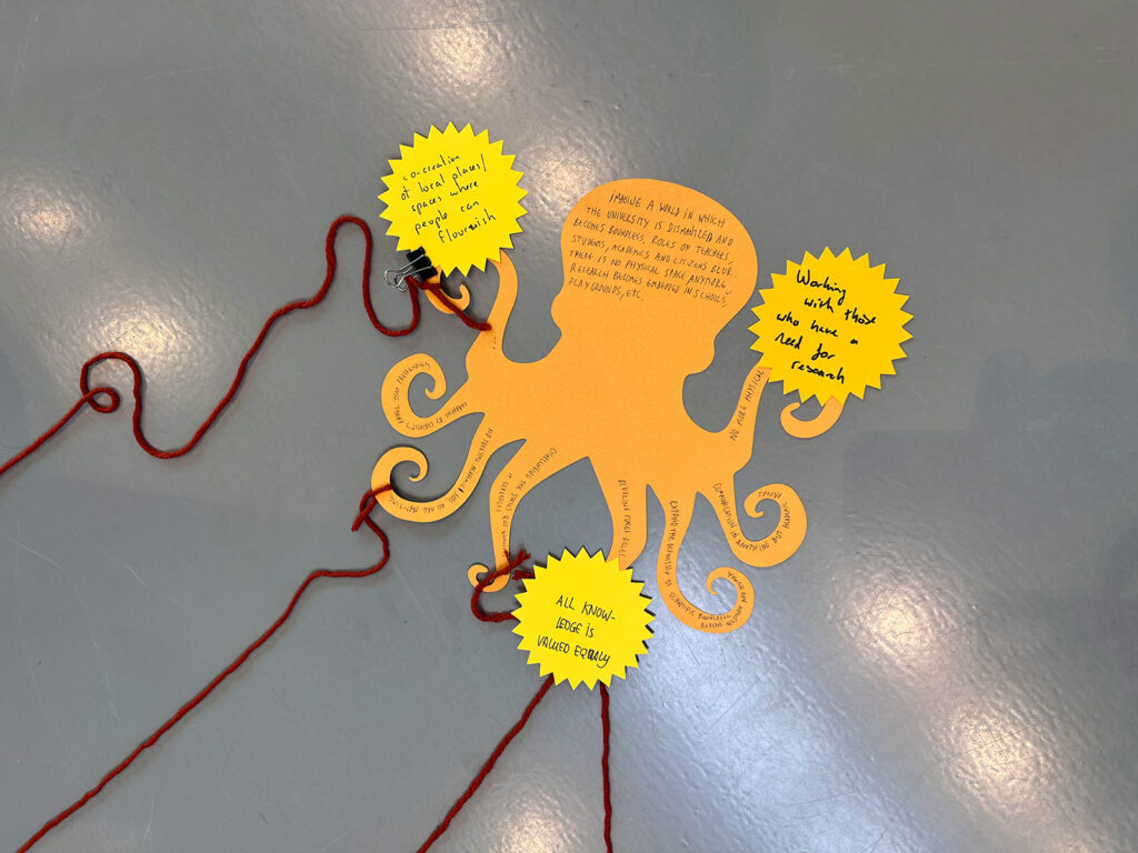 A yellow paper octopus with handwritten notes on its body and sun-shaped cutouts, connected to red yarn strands on a gray surface. The notes mention friendship and making friends.