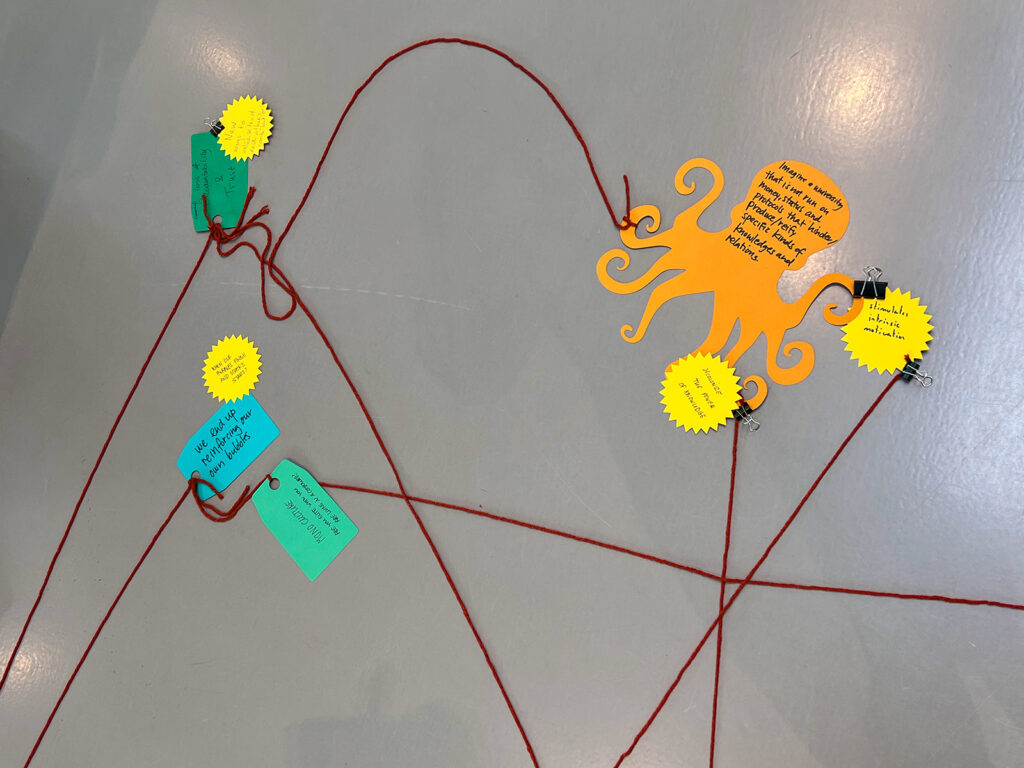 Red strings connect colorful paper notes, including an orange octopus-shaped note with writing, on a gray surface. Tags and yellow starbursts with handwritten messages are clipped to the strings.