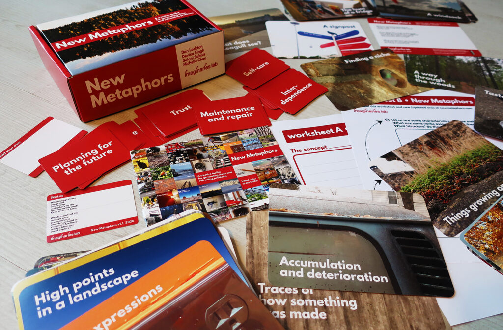A tabletop displays the "New Metaphors" card game set. The open box and various colorful cards with text and images are spread out, showcasing categories like "Planning for the future," "worksheet A," and landscape photos.
