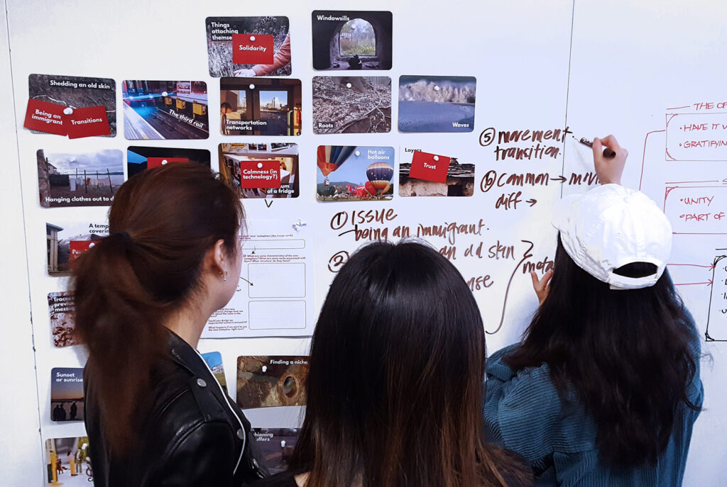Three people stand facing a whiteboard covered with notes, sketches, and photos. One person writes with a marker while the others observe and discuss the ideas displayed.