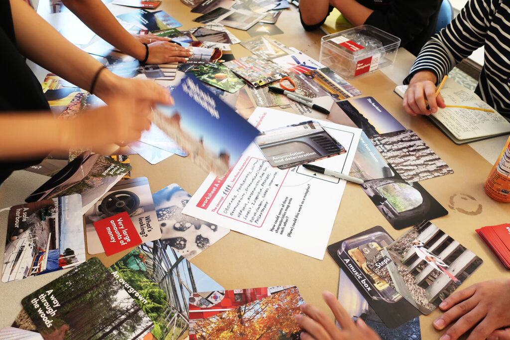 Several people sit at a table covered with assorted cards featuring photos and text, as well as notebooks, pens, and a drink can, suggesting a group activity or brainstorming session.