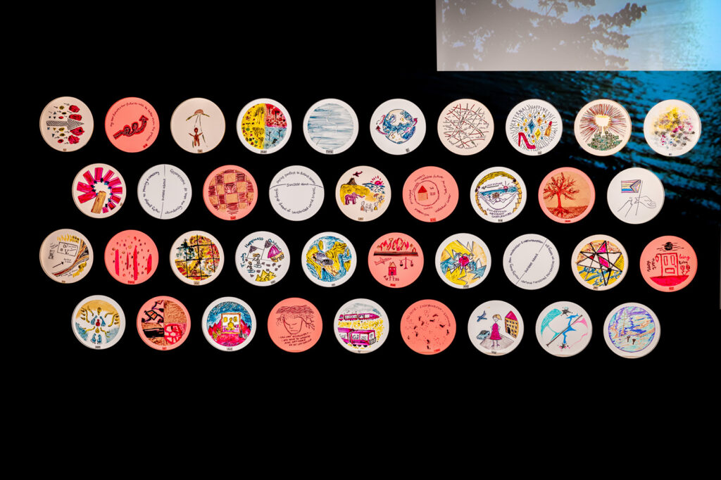 A grid of colorful, round coasters with various hand-drawn illustrations and patterns is arranged against a black background; a small portion of a blue, leafy image appears in the top right corner.