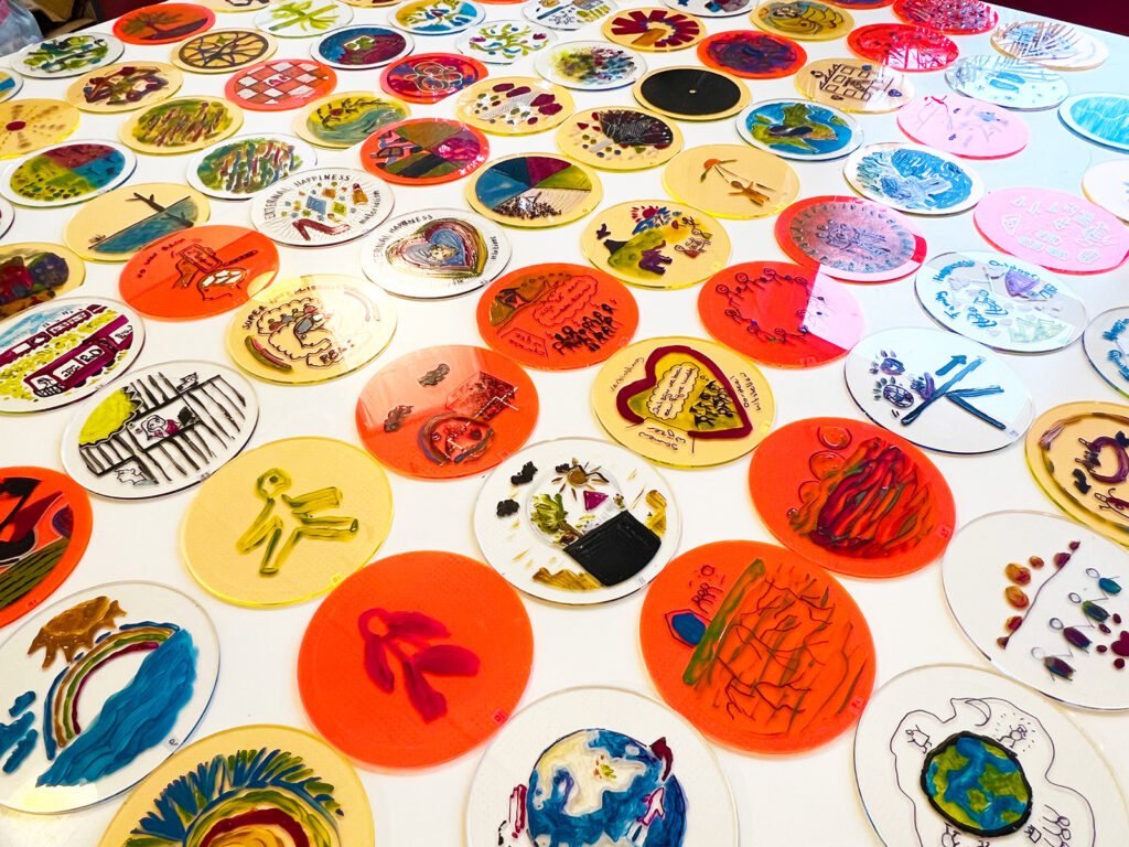 A table covered with numerous round, colorful artworks on clear plastic discs, each featuring unique, hand-drawn designs, patterns, and illustrations in various colors.