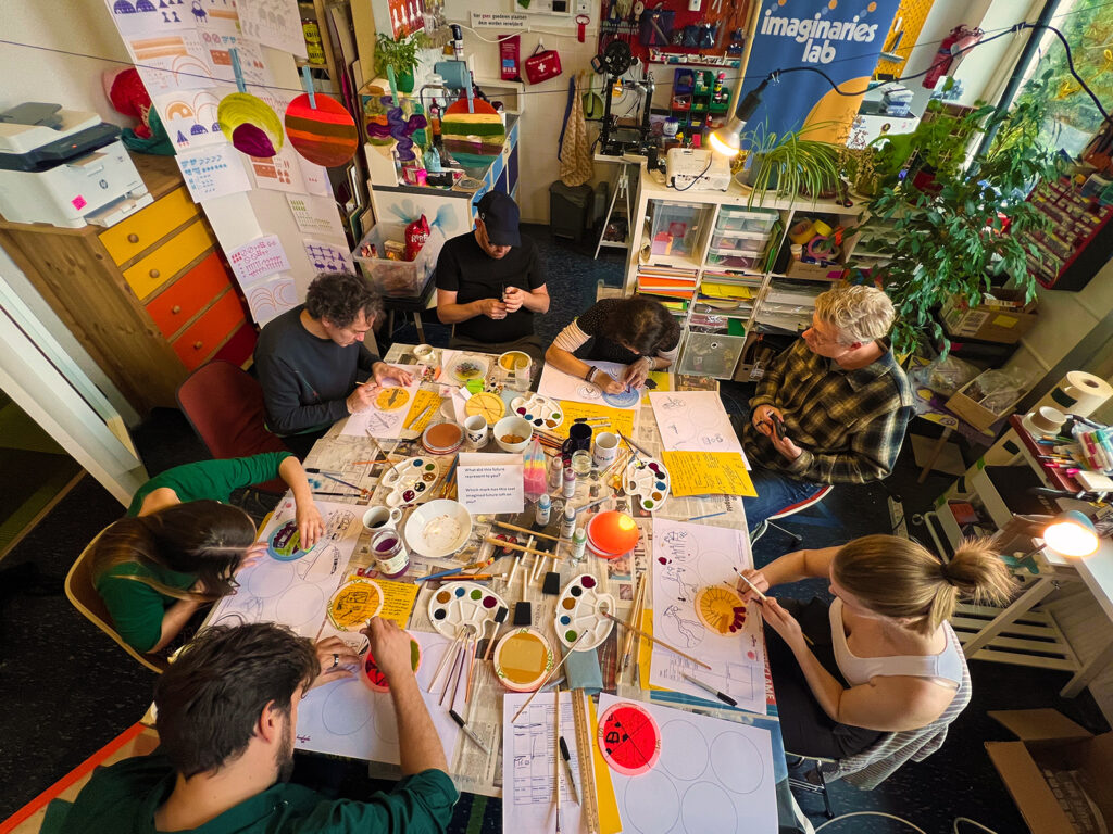 Seven people sit around a cluttered table filled with papers, paints, and art supplies, working on creative projects in a colorful, cozy studio decorated with rainbow items and a sign reading "imaginaires lab.