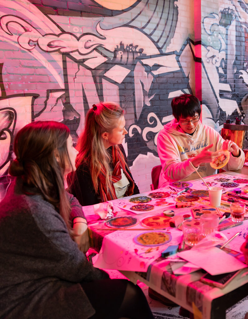 Three people sit around a table covered with art supplies, engaging in a creative activity. They are indoors, and a colorful mural covers the brick wall behind them. The lighting has a pinkish hue.