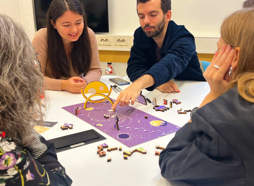 Four people sit around a table playing a colorful tabletop board game. One man points at the game board while others watch and discuss, appearing engaged and focused. The setting is a bright room with white walls.