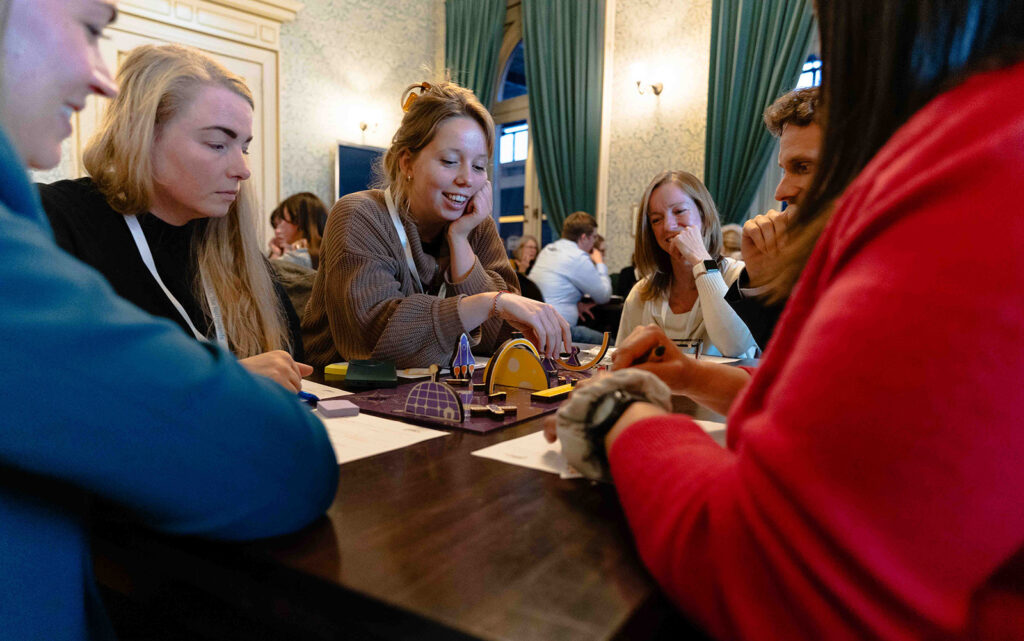 A group of people sit around a table, smiling and interacting with a board game or activity. Papers are on the table, and other groups are visible in the background of the warmly lit room.