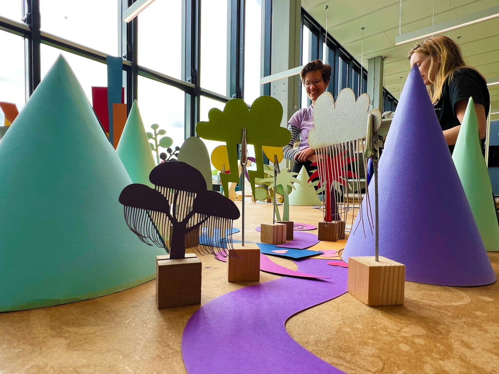 Two people work on a colorful paper diorama featuring cone-shaped mountains, trees, and a winding purple path on a wooden table near large windows in a bright room.