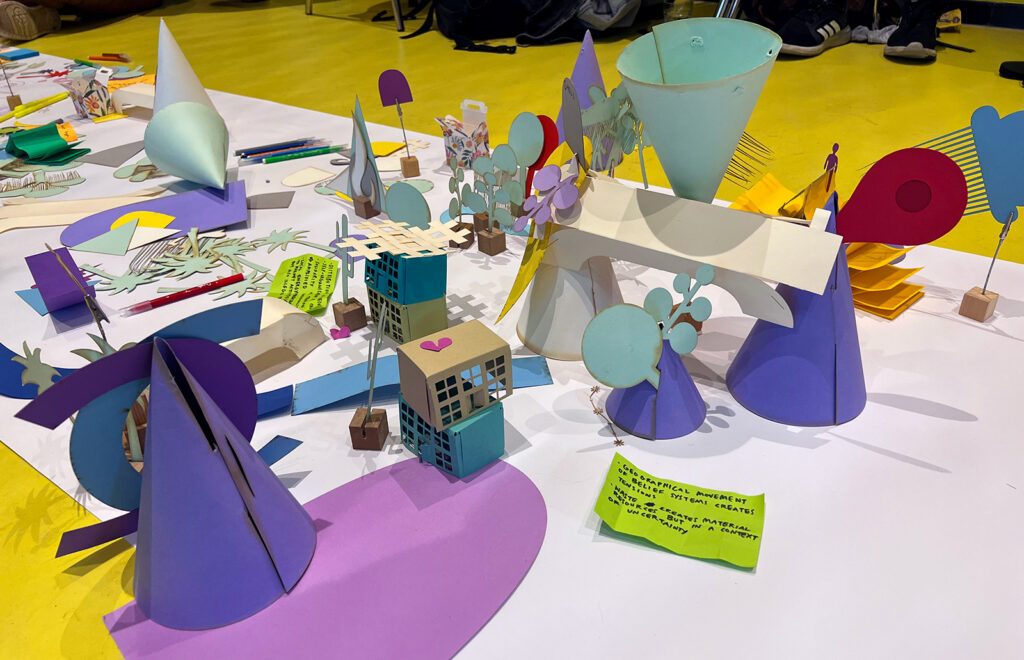 A colorful tabletop display features abstract paper models of buildings, trees, and cones, with handwritten notes on bright paper, all arranged on a white surface over a yellow floor.